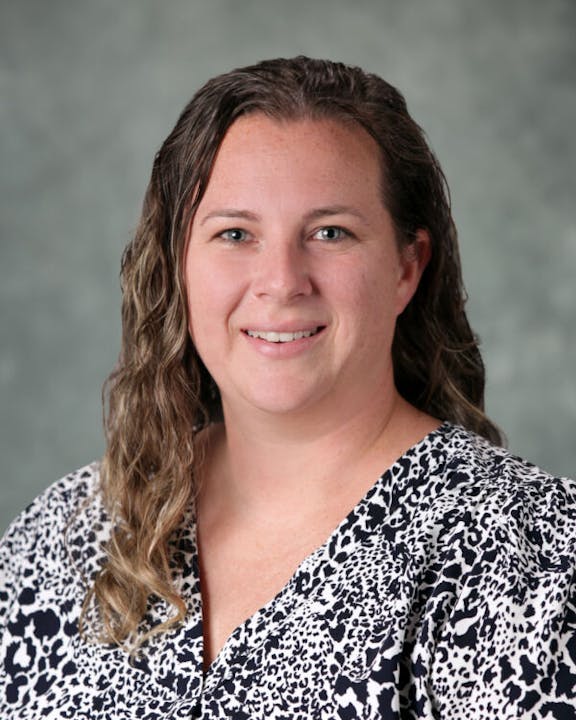 A person with wavy hair wearing a patterned top, smiling against a gray background.
