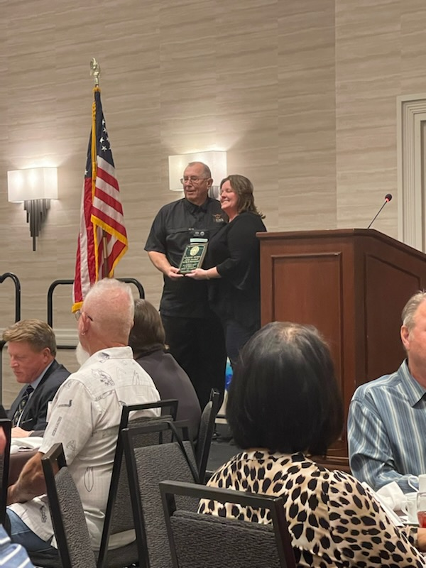 A man and woman stand near a podium holding a plaque, with people seated at tables and an American flag in the background.