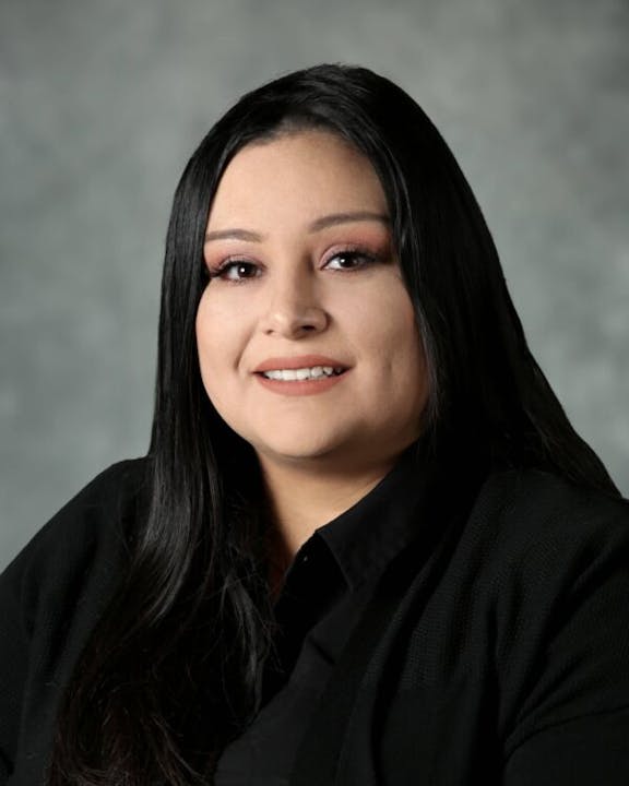 A person with long dark hair wearing a black shirt, smiling against a gray background.