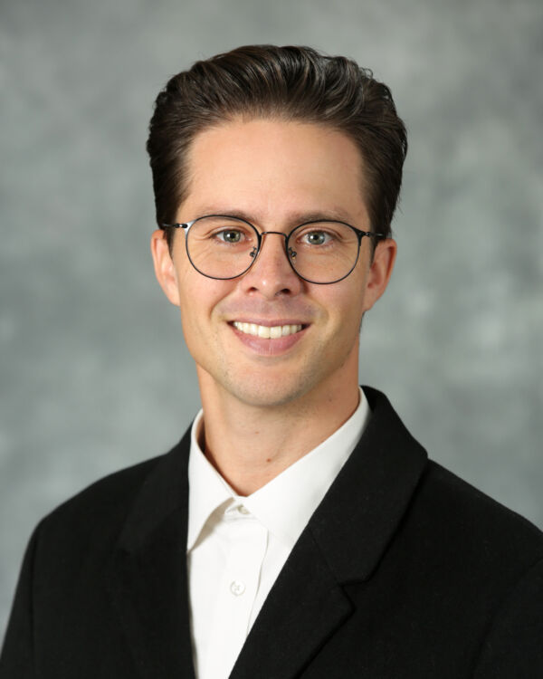 A person wearing glasses and a suit, smiling against a neutral background.