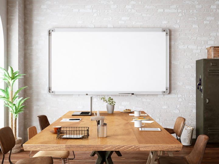 A modern office setting with a wooden table, chairs, a large whiteboard, plant, lockers, and office supplies on the table.