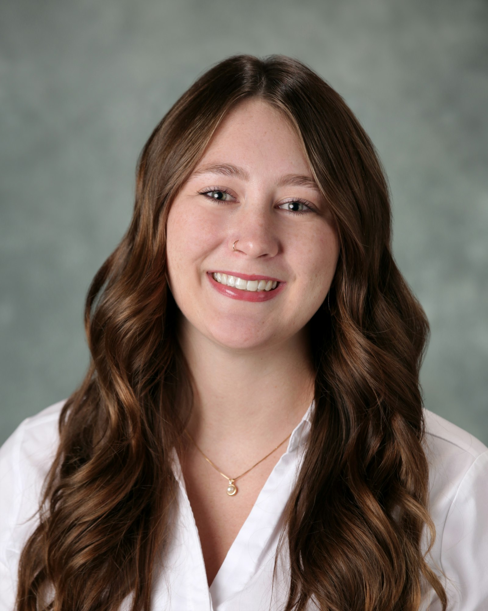 A woman with long, wavy hair smiles, wearing a white shirt and a necklace, against a soft, blurred background.