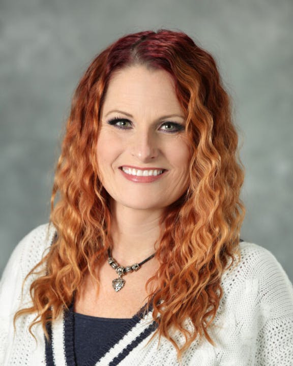 A person with wavy red hair, smiling, wearing a white sweater and a necklace.