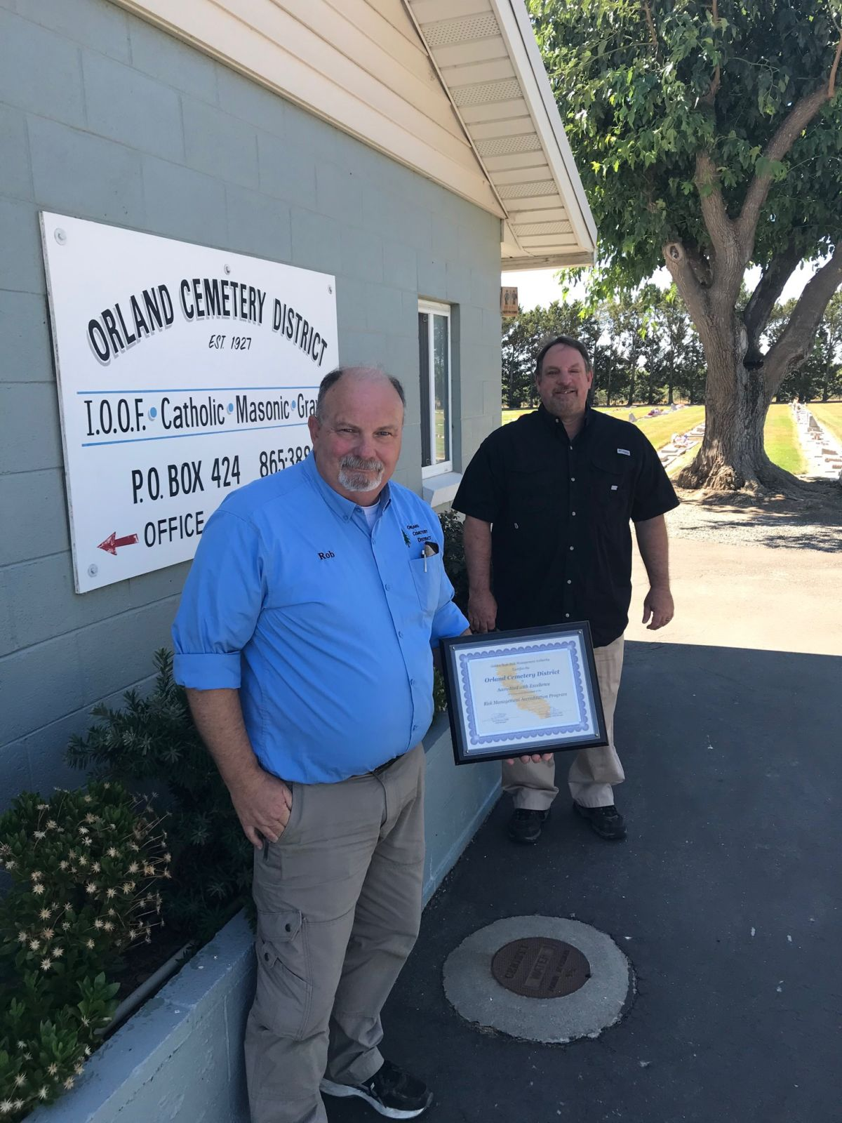Two men stand outside Orland Cemetery District office, one holding a framed certificate.