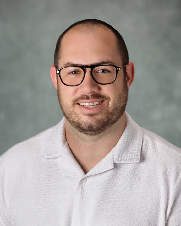 This image features a man with glasses smiling, wearing a light-colored shirt, against a soft, neutral background.