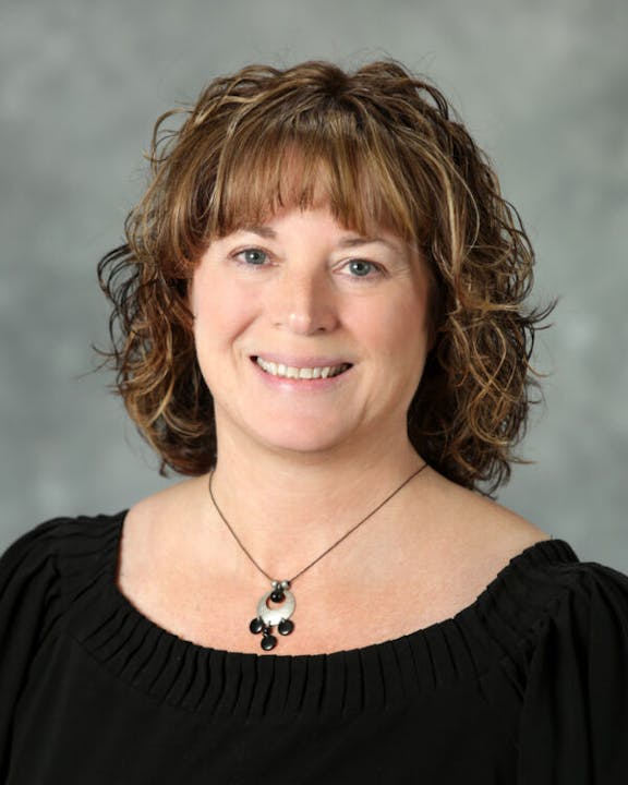 A person with curly hair smiling, wearing a black top and a necklace.