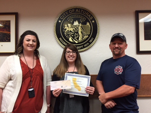 Three people standing, one holding a certificate and check, in front of a "City of Alturas, California" seal.