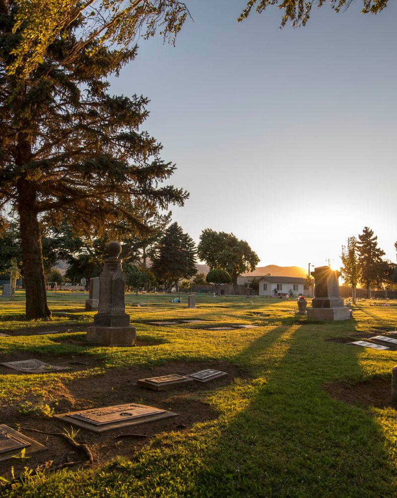 A cemetery at sunset, with gravestones, trees, and a small building in the background.