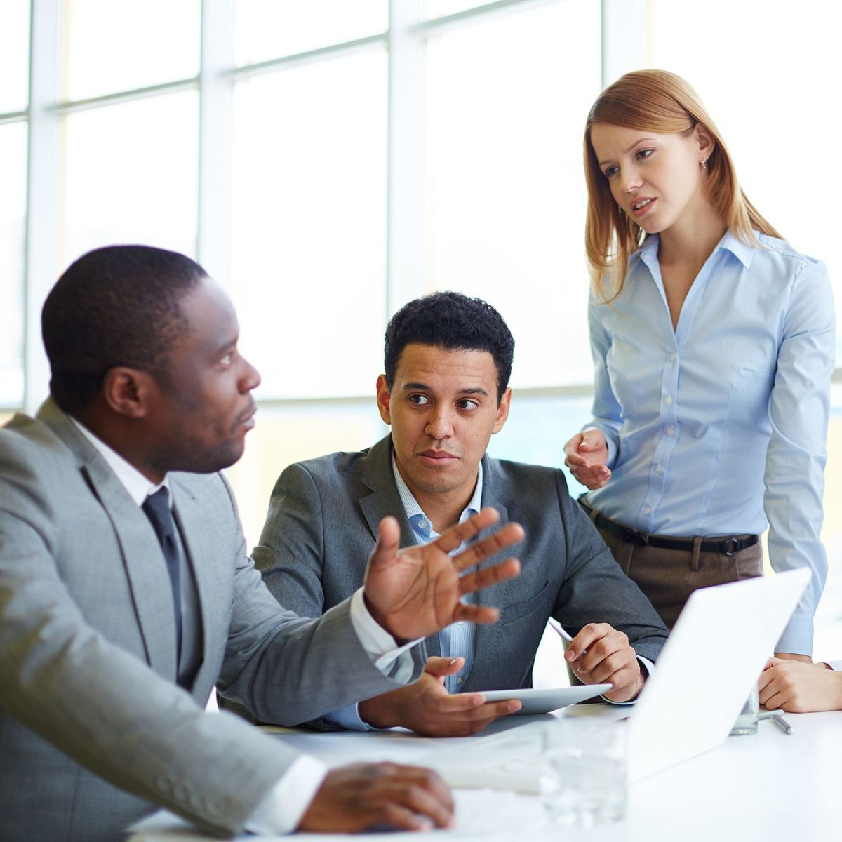Three people in a business meeting, two men sitting with a laptop and tablet, one woman standing, all in a discussion.
