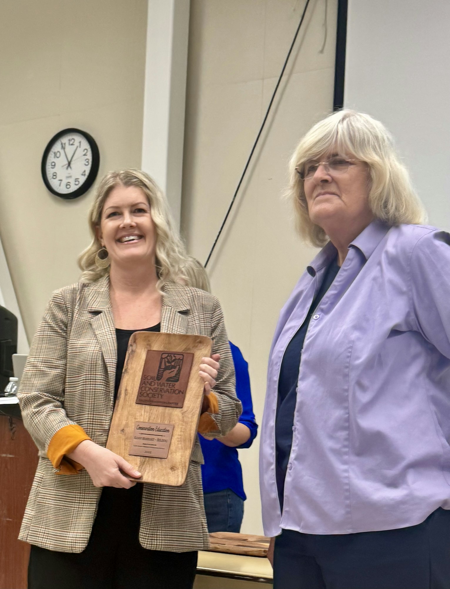 Two women stand together, one holding an award plaque from the Soil and Water Conservation Society, celebrating an achievement.