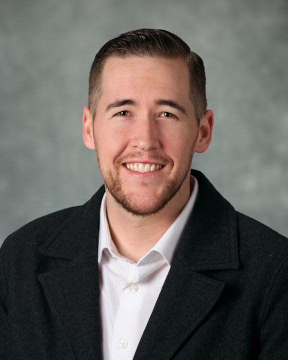 A man in a dark suit jacket and white shirt smiling against a gray background.