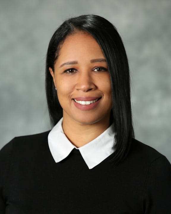A woman with long black hair, wearing a black top with a white collar, smiling against a gray background.