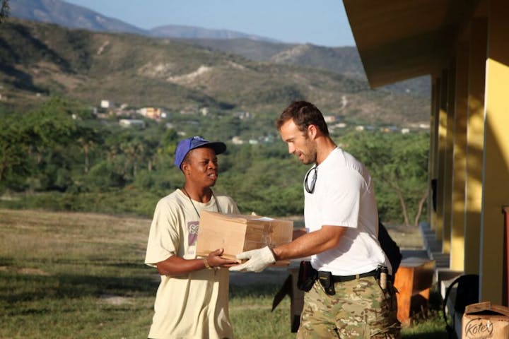 Two people exchanging a box outdoors with mountains in the background.
