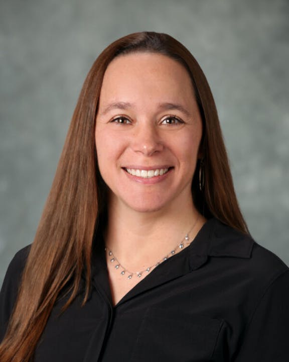 A person with long brown hair smiling, wearing a black top and a necklace, against a gray background.