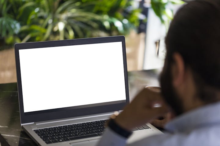 A person looking at a laptop with a blank white screen, set against a backdrop of green plants.