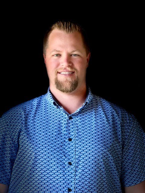 A person in a blue patterned shirt smiling against a black background.