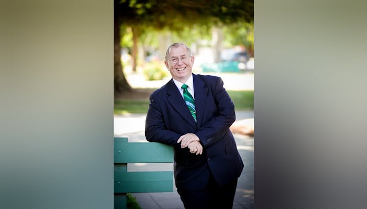 A man in a suit with a green tie is smiling, leaning on a green fence in a sunny park.