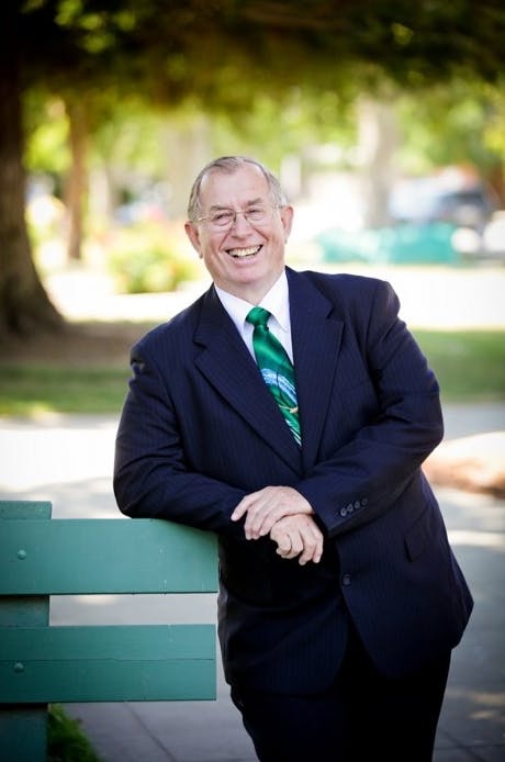 A man in a suit with a green tie is smiling, leaning on a green fence in a sunny park.