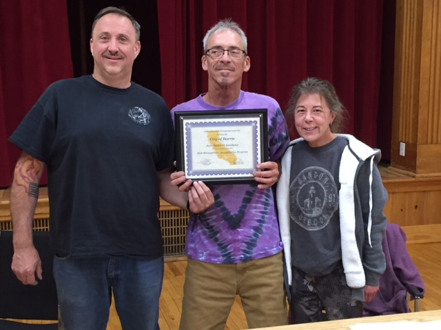 Three people standing indoors; the middle person holds a framed certificate.