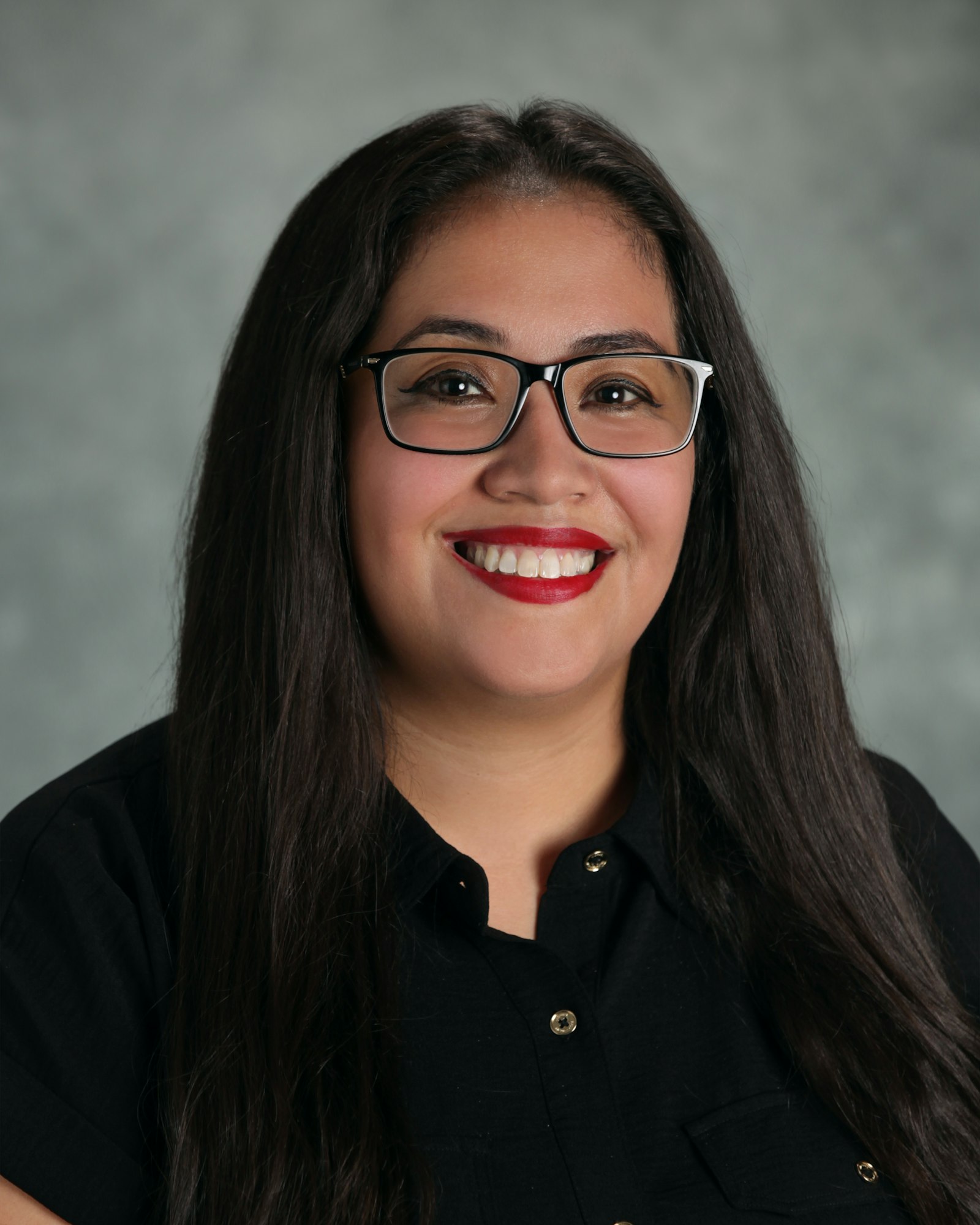 The image shows a woman with long dark hair, wearing glasses and a black shirt, smiling against a neutral background.