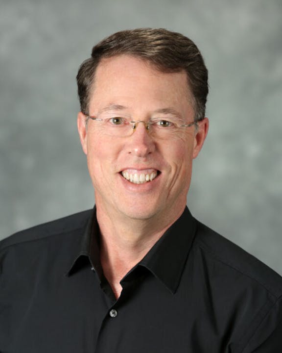 A smiling person with glasses wearing a black shirt, against a gray background.