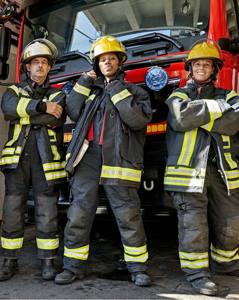 Three firefighters in protective gear stand confidently in front of a red fire truck.