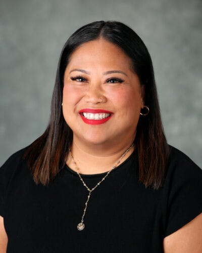 A person smiling, wearing a black shirt and silver necklace, with a gray background.