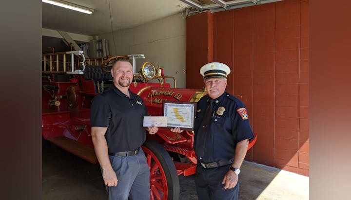 Two men stand in front of an antique fire truck, holding a certificate and a check.