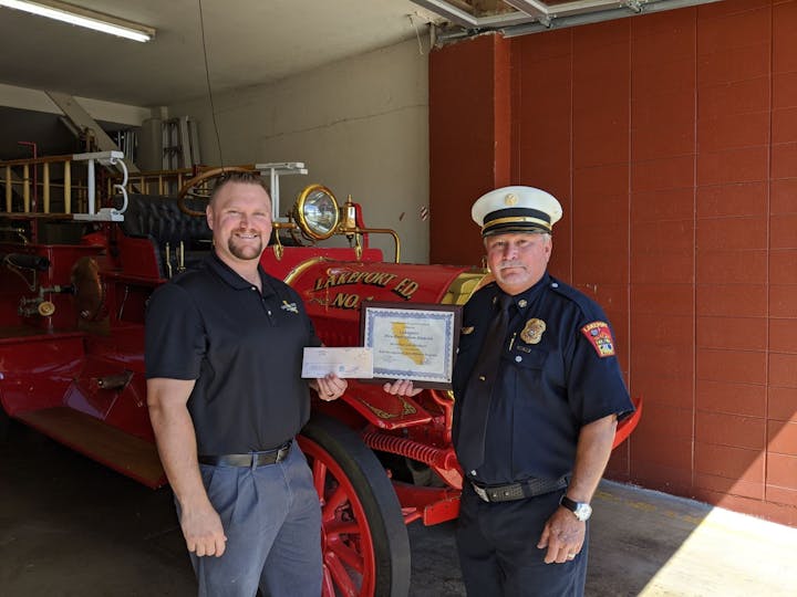 Two men stand in front of an antique fire truck, holding a certificate and a check.