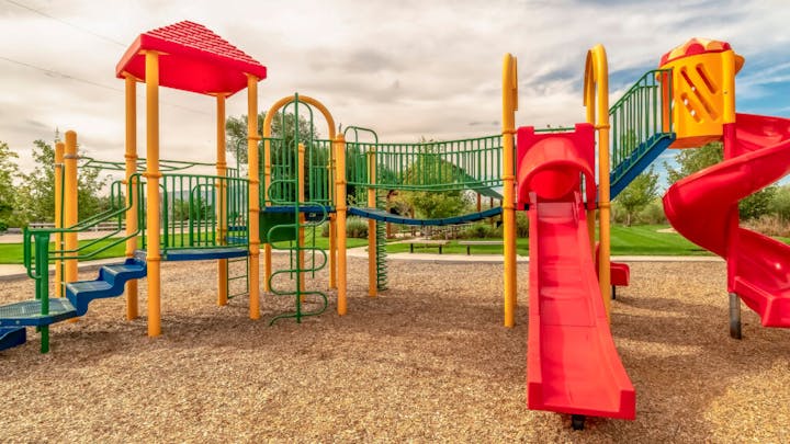 A colorful playground with slides, climbing structures, and wood chip ground cover, set in a park with trees.