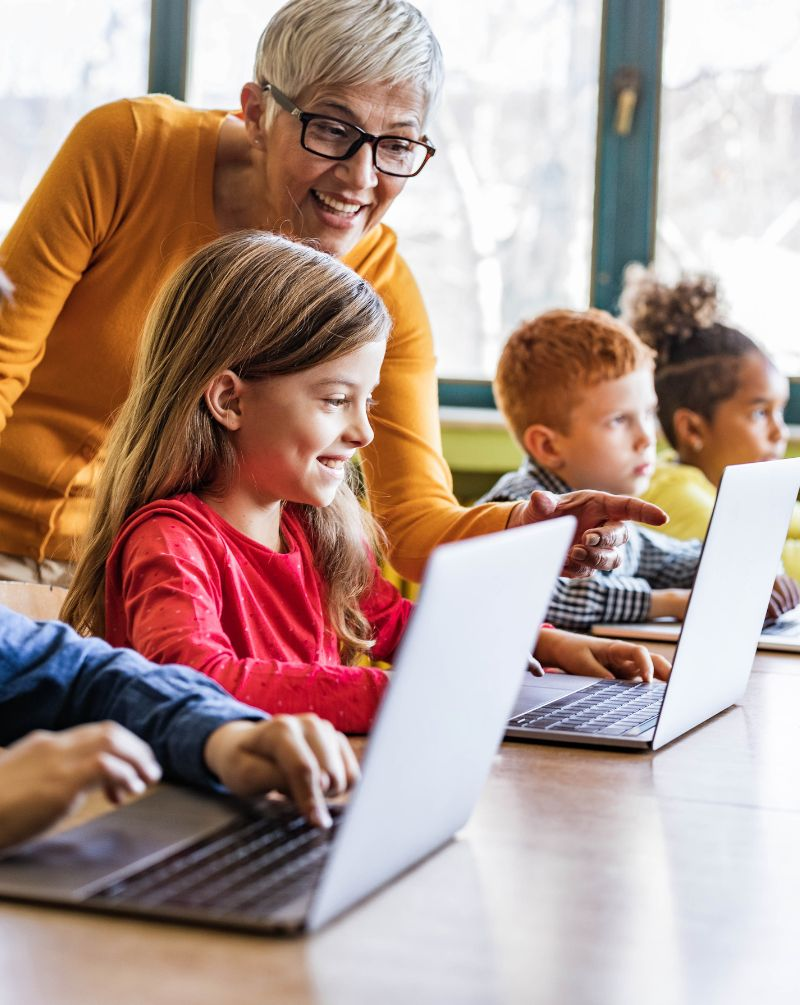 A teacher helps young students working on laptops in a classroom setting.