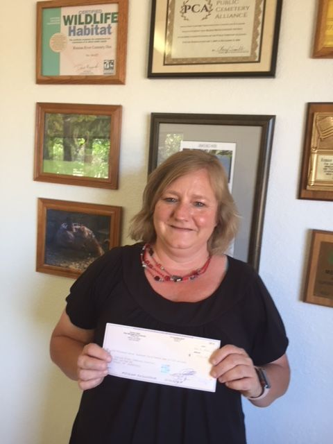 A woman holding a check, standing in front of a wall with framed certificates and pictures.