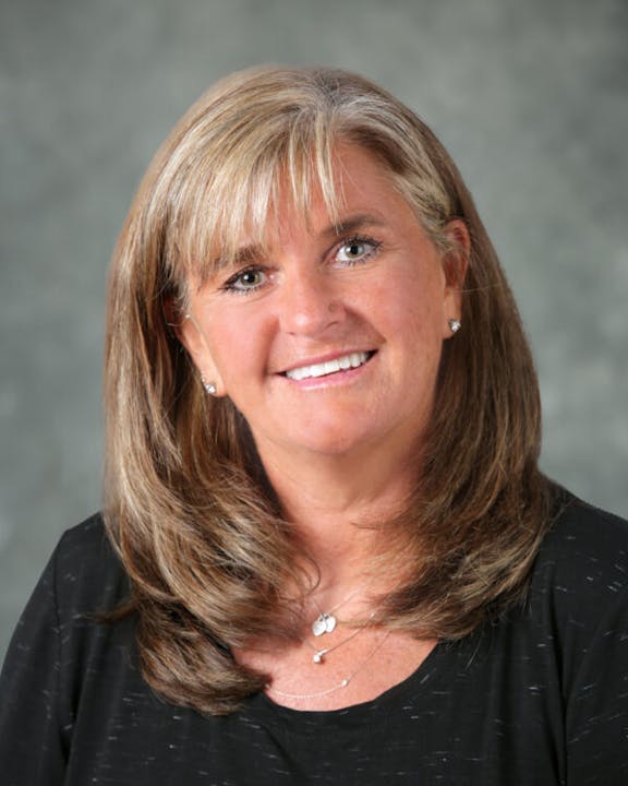 A person smiling, with shoulder-length brown hair, wears a black top and necklace, set against a gray background.