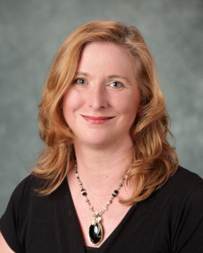 A person with shoulder-length hair, wearing a black top and necklace, against a neutral background.