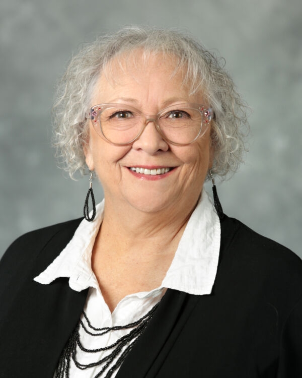 An older woman with curly gray hair, glasses, and a smile, wearing a white blouse and black jacket with dangly earrings.