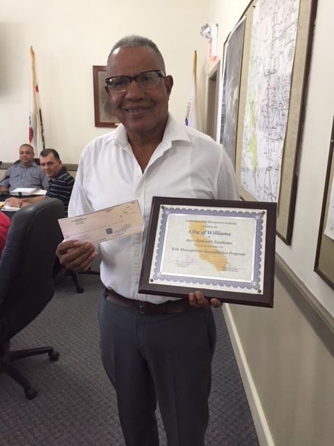 A man holding a certificate and a check, smiling in a room with maps on the wall.