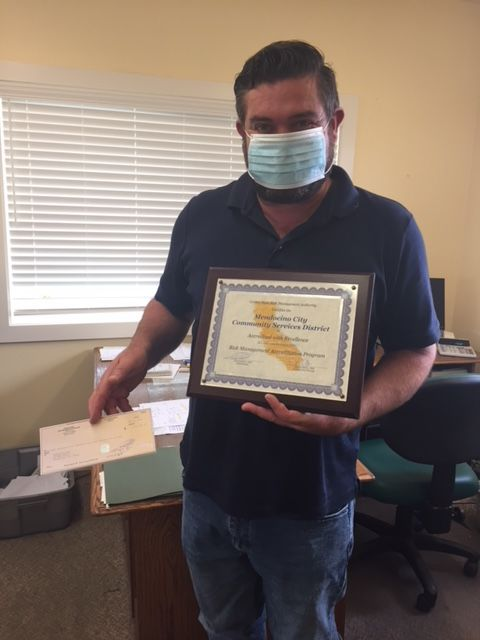 A person wearing a mask holds a framed certificate and an envelope in an office setting.