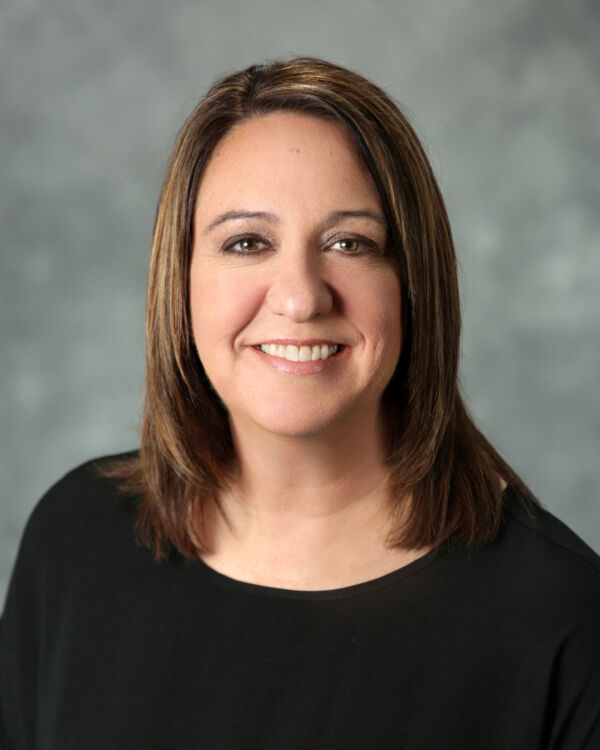 A woman with shoulder-length brown hair smiling, wearing a black top, against a gray background.