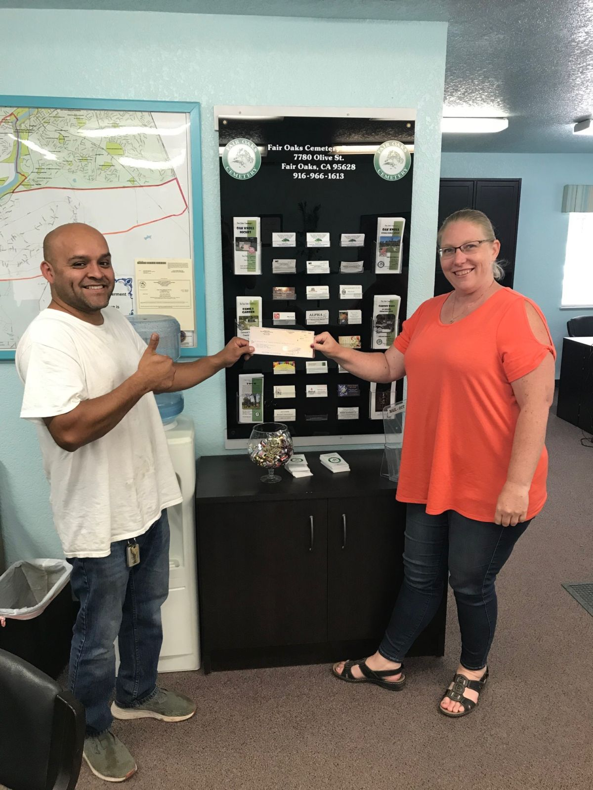 Two people smiling while holding a check in an office with brochures displayed on the wall.