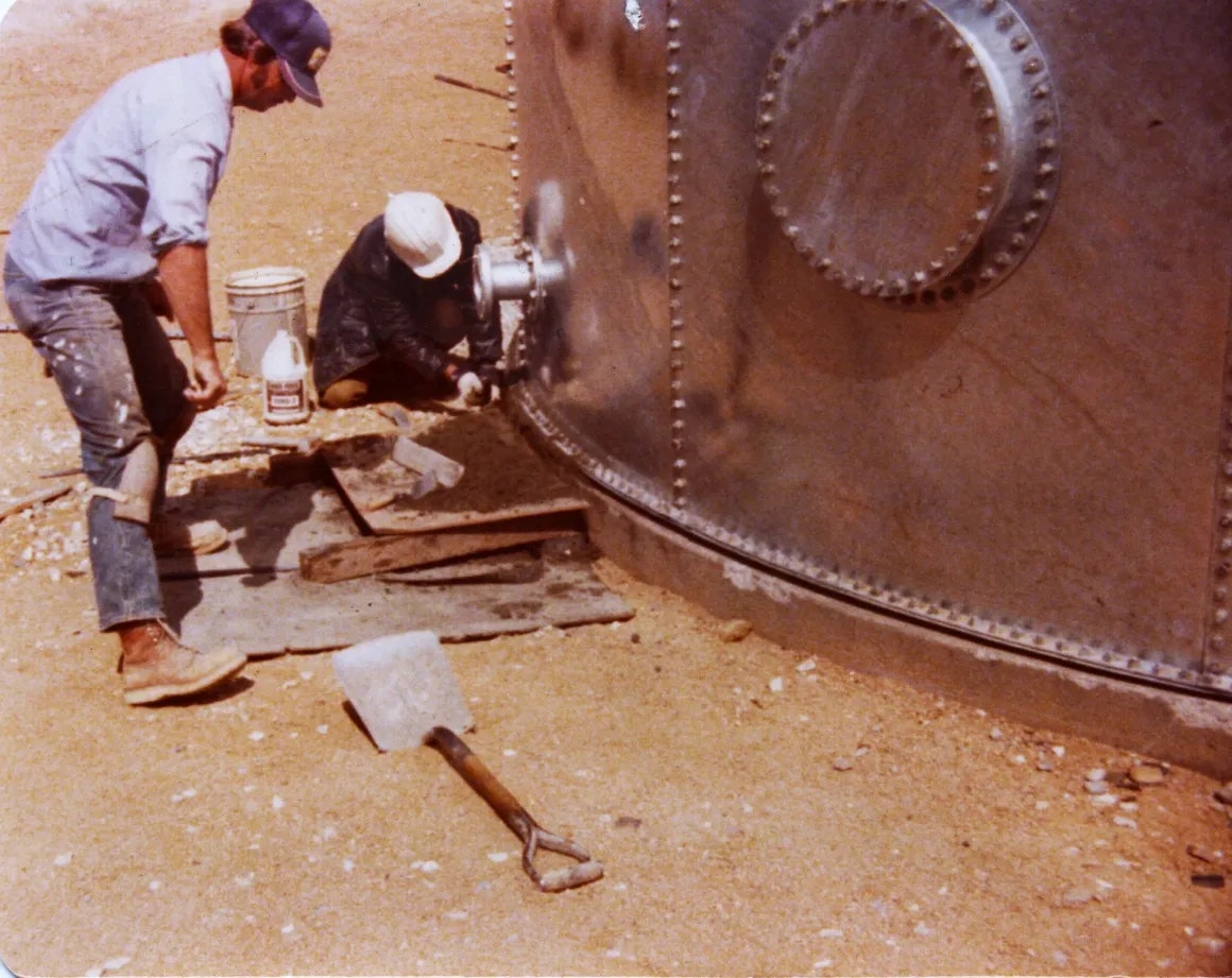 Two people repairing a large metal container outdoors, with tools on the ground.