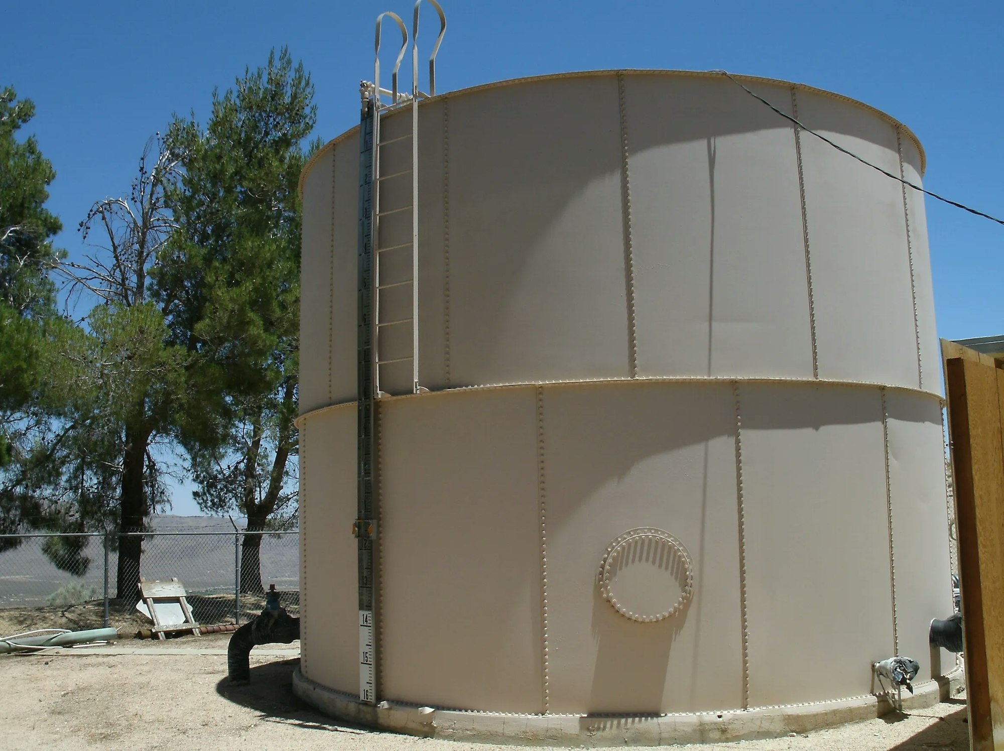 Large outdoor water storage tank with a ladder, in a fenced area with trees in the background.