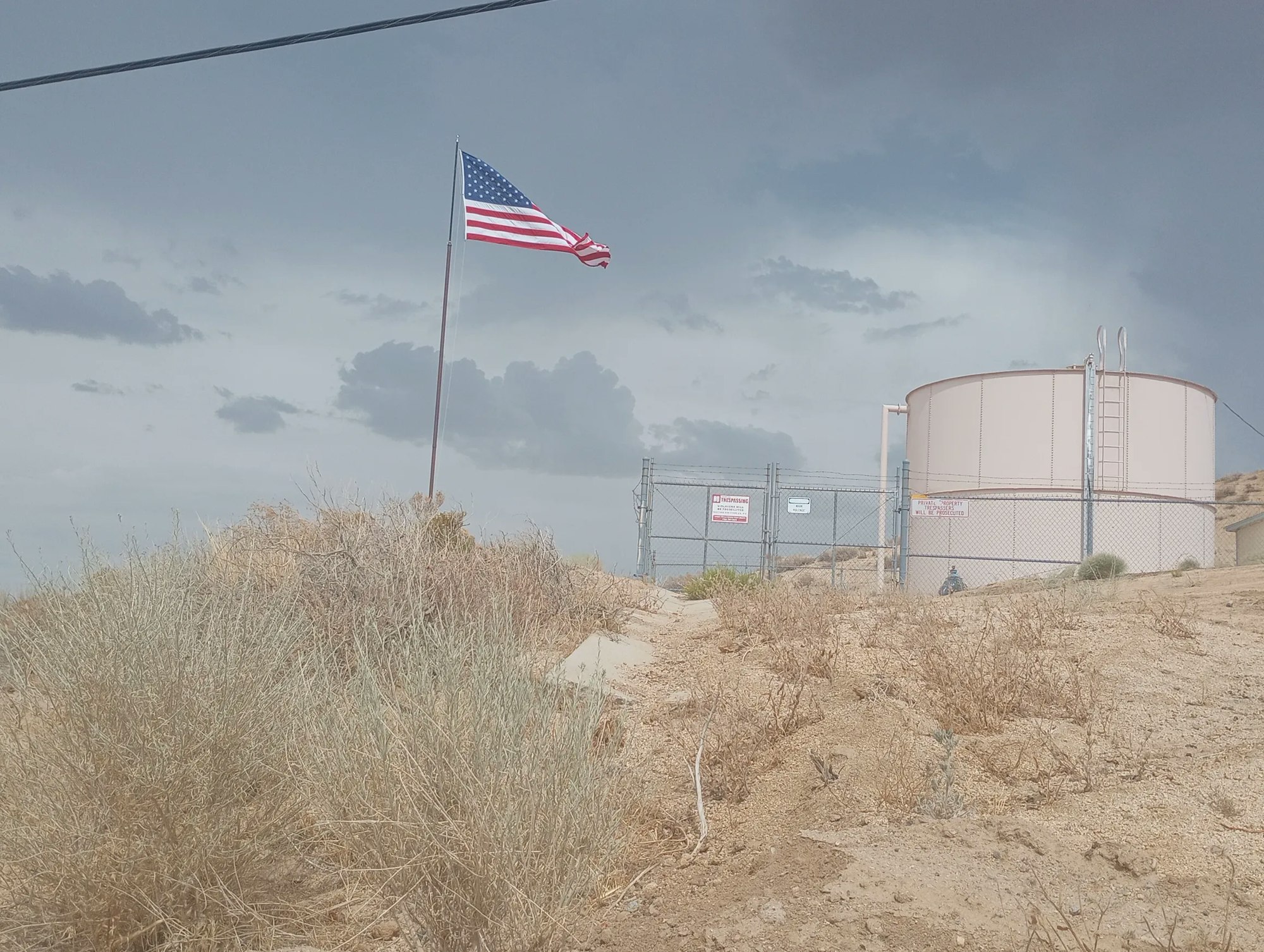 American flag waving, stormy sky, fenced storage tanks, and dry vegetation.