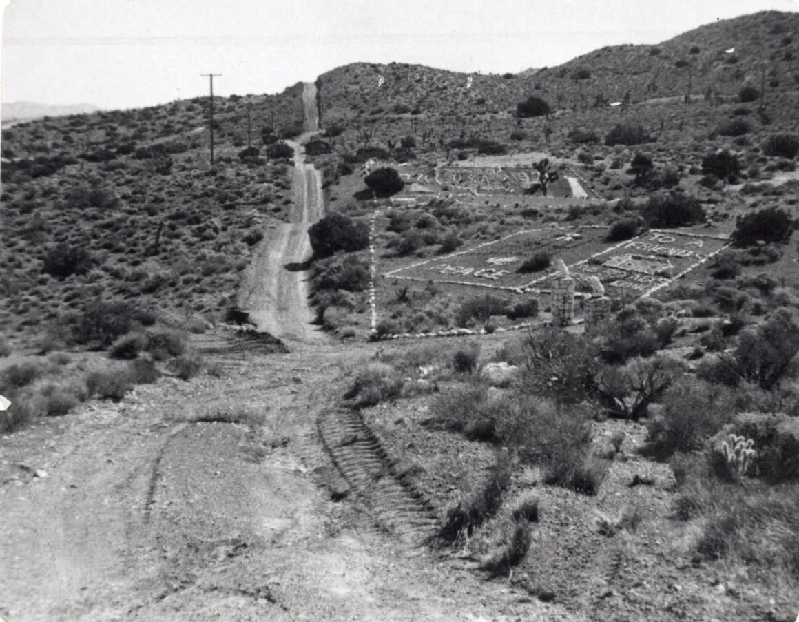 A black and white photo of a rugged landscape with a dirt road, some vegetation, and a patterned layout in the distance.