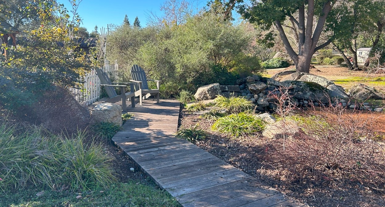 A garden scene with a wooden path, two Adirondack chairs, greenery, rocks, and trees under a clear blue sky.