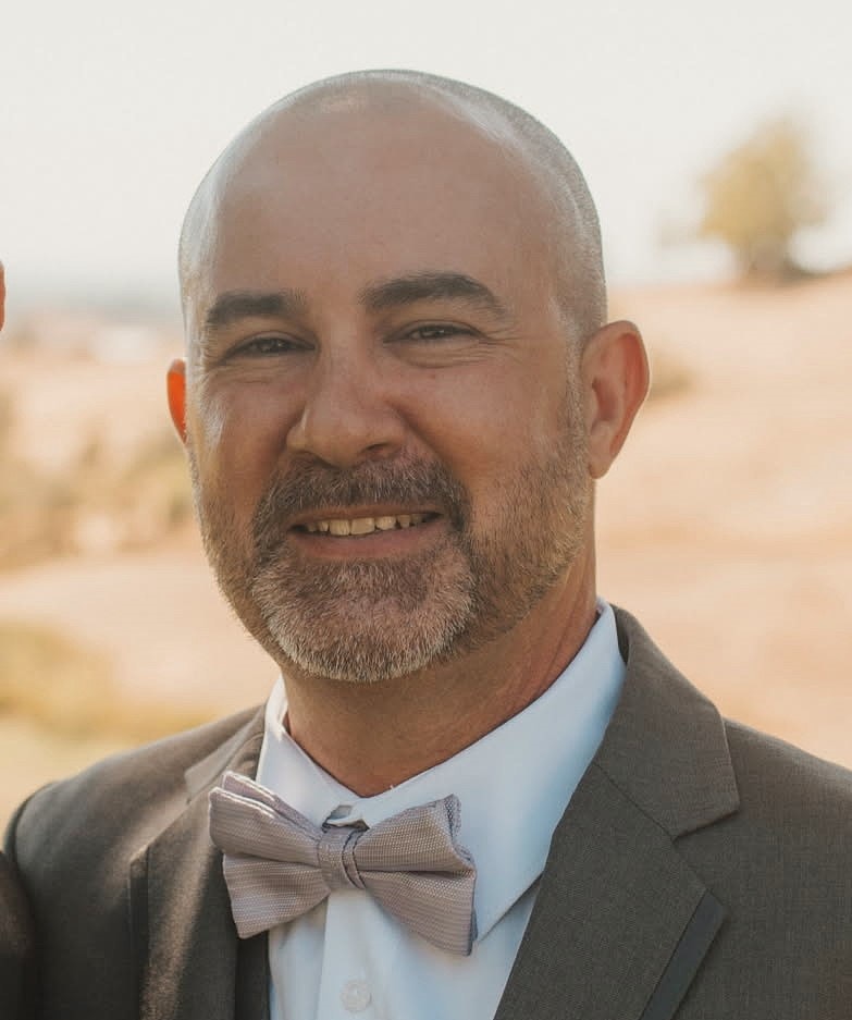 A man smiling, wearing a suit with a bow tie, in an outdoor setting.