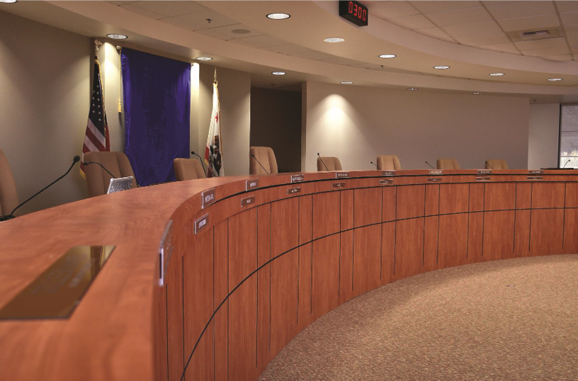 Empty council chamber with wooden desks, microphones, and nameplates; American and state flags in the background.
