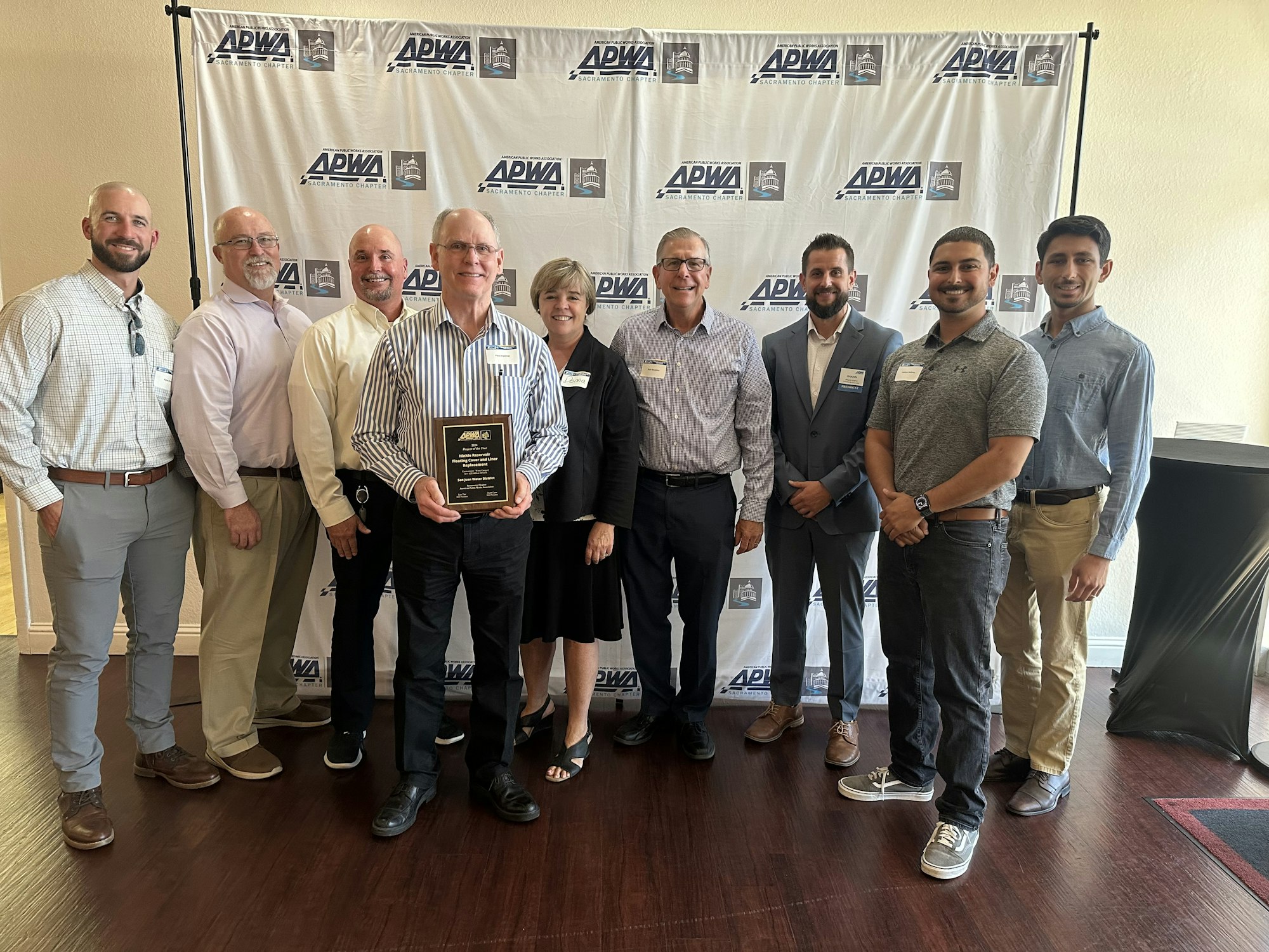 A group of eight people in business casual attire, holding an award, posing before a banner with logos.
