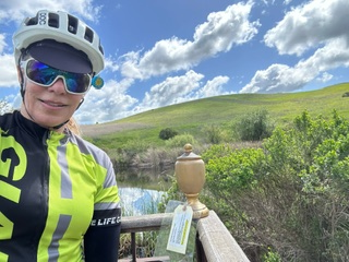 A cyclist with helmet and sunglasses next to a trophy on a railing, greenery and clouds in the background.