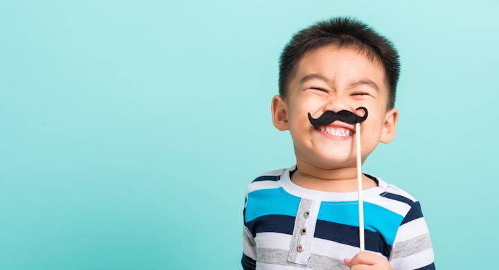 A smiling child holds a fake mustache on a stick against a light blue background, showcasing a playful expression.