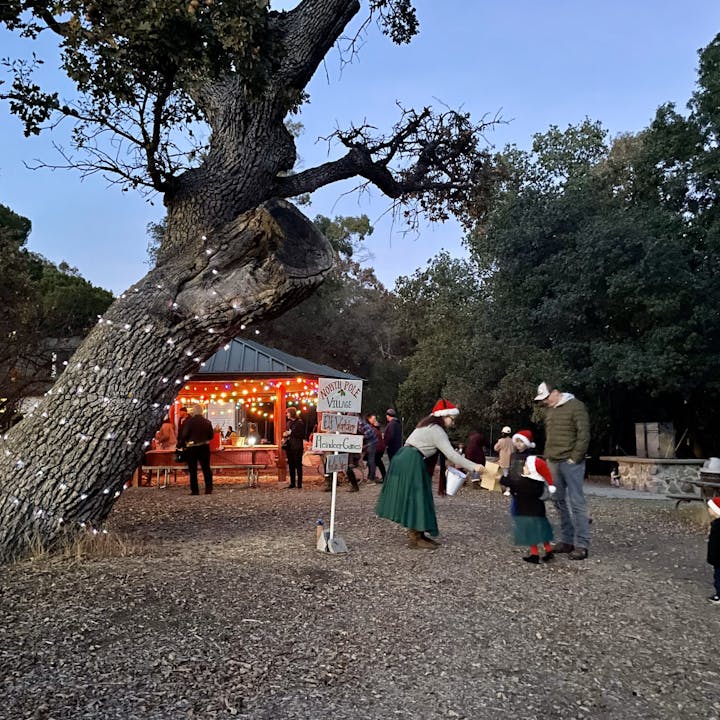 A festive scene at a holiday event with lights, a sign for "North Pole Village," and people in winter attire, including Santa hats.